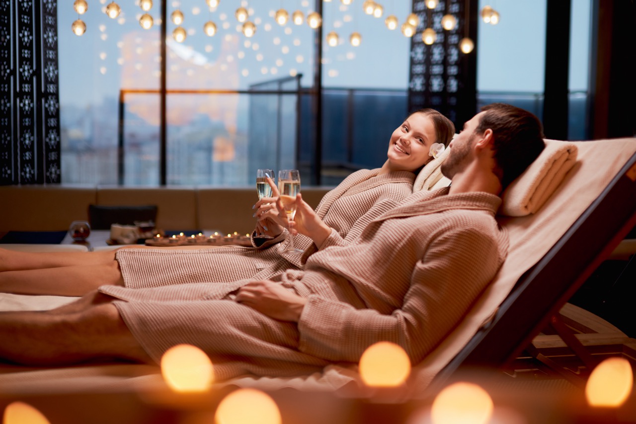 Married couple together relaxing in spa salon, lying on beds drinking champagne, using candles